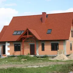 Dom w budowie z czerwonym dachem, gotowy do ekspertyzy technicznej budynku. Wokół materiały budowlane i piasek.