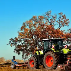 Zielony traktor z pługiem pod jesiennym drzewem, przygotowujący pole do odrolnienia działki.