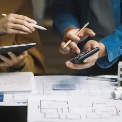Architekci analizują plany, liczą koszty i omawiają, ile kosztuje odbiór domu. Na stole szkice, tablet, kalkulator i model budynku.