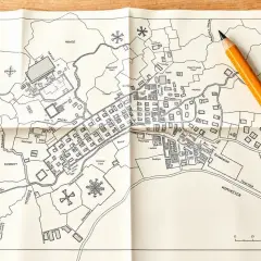 Składana mapa zasadnicza z zaznaczonymi budynkami, drogami i rzeką. Obok leży żółty ołówek, gotowy do naniesienia kolejnych szczegółów.