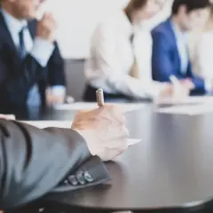 Ludzie w garniturach przy stole, jeden z nich pisze. Wszyscy czekają na kworum, by rozpocząć ważne spotkanie.