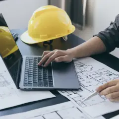 Architekt analizuje plany, obliczając powierzchnię całkowitą budynku. Na biurku leży żółty kask i laptop.