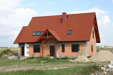Dom w budowie z czerwonym dachem, gotowy do ekspertyzy technicznej budynku. Wokół materiały budowlane i piasek.