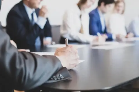 Ludzie w garniturach przy stole, jeden z nich pisze. Wszyscy czekają na kworum, by rozpocząć ważne spotkanie.