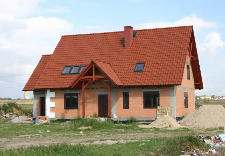 Dom w budowie z czerwonym dachem, gotowy do ekspertyzy technicznej budynku. Wokół materiały budowlane i piasek.