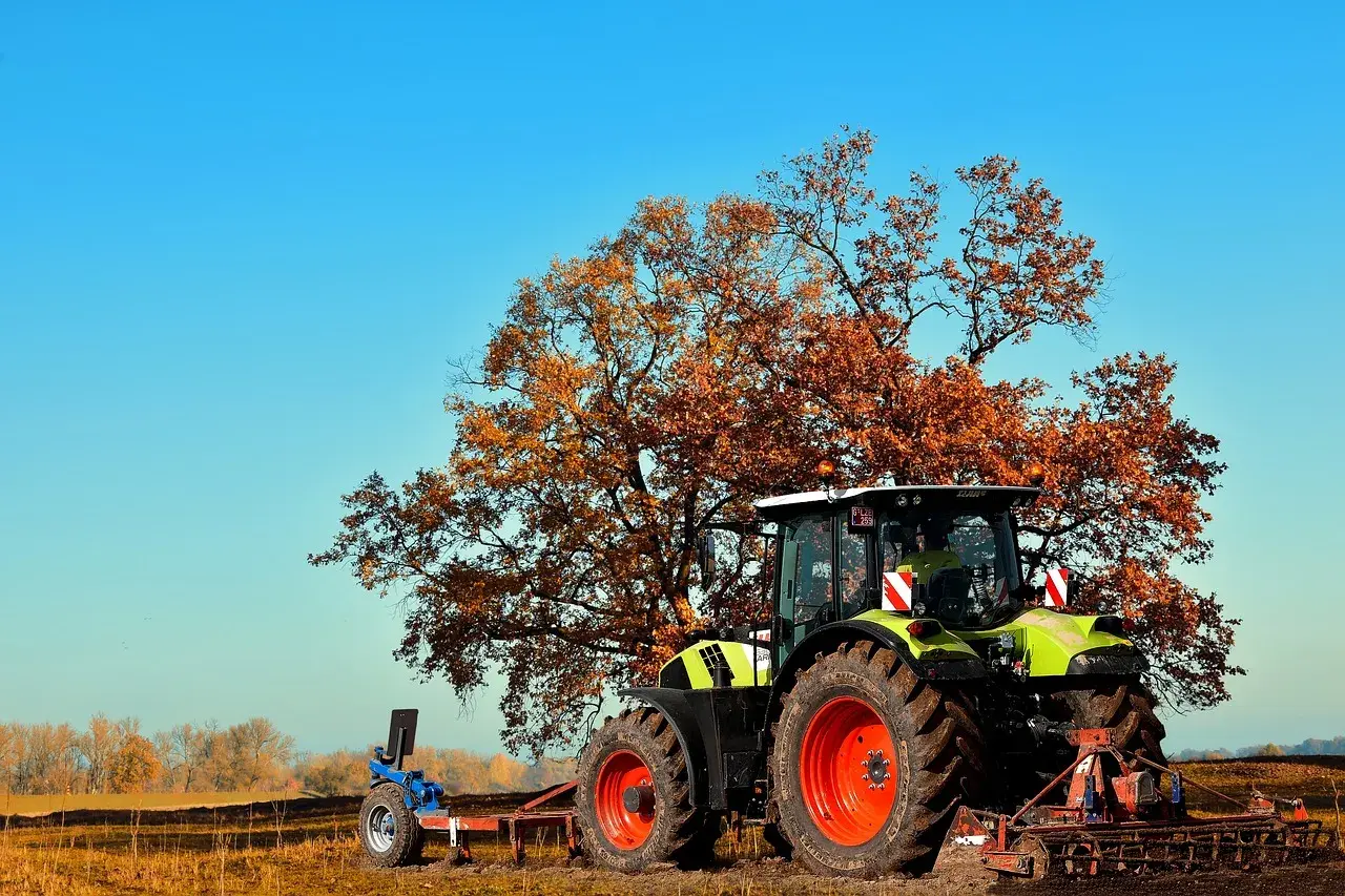 Zielony traktor z pługiem pod jesiennym drzewem, przygotowujący pole do odrolnienia działki.