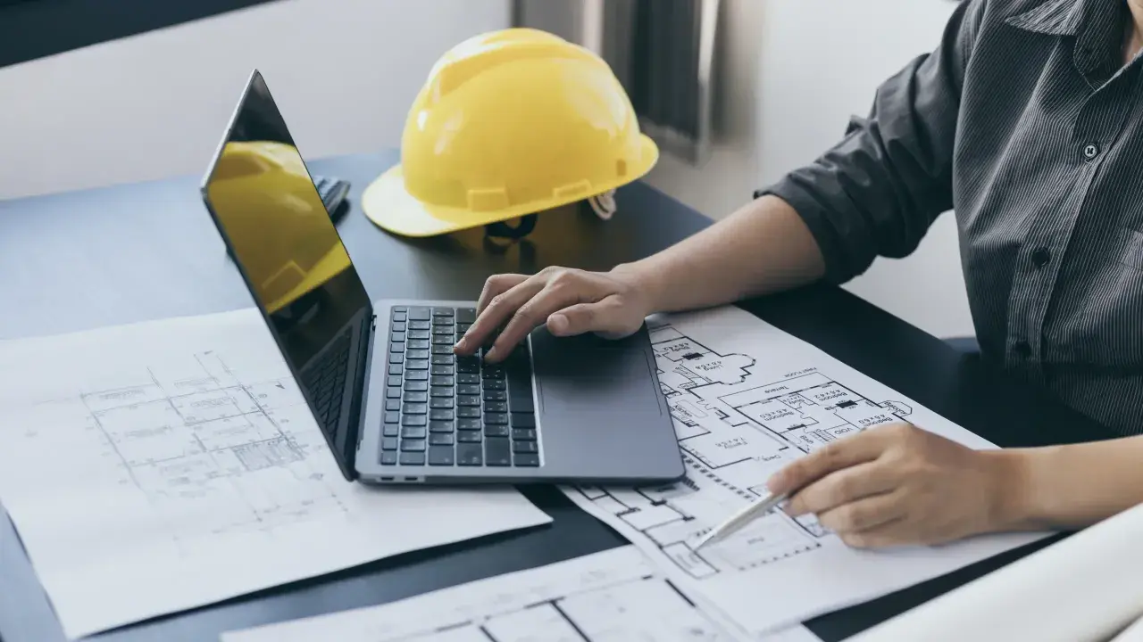 Architekt analizuje plany, obliczając powierzchnię całkowitą budynku. Na biurku leży żółty kask i laptop.