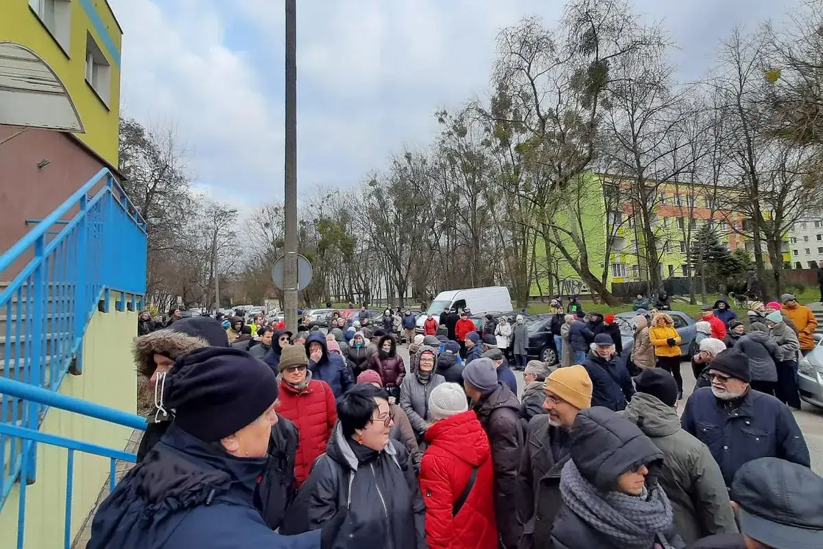 Tłum ludzi na zewnątrz, być może czekających na spotkanie rady nadzorczej. W tle budynki i drzewa.
